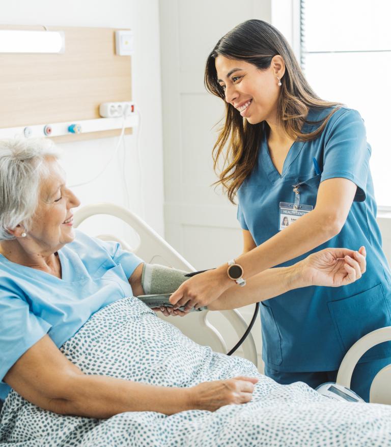 Nurse talking to senior woman in hospital bed. She is measuring her blood pressure.